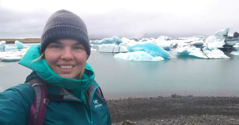 Iemand met een grijze muts en een blauwgroen jasje glimlacht naar de camera, met achter zich ijsbergen die in een koud, grijs meer drijven. Een rotsachtige oever en een bewolkte hemel maken het tafereel compleet.